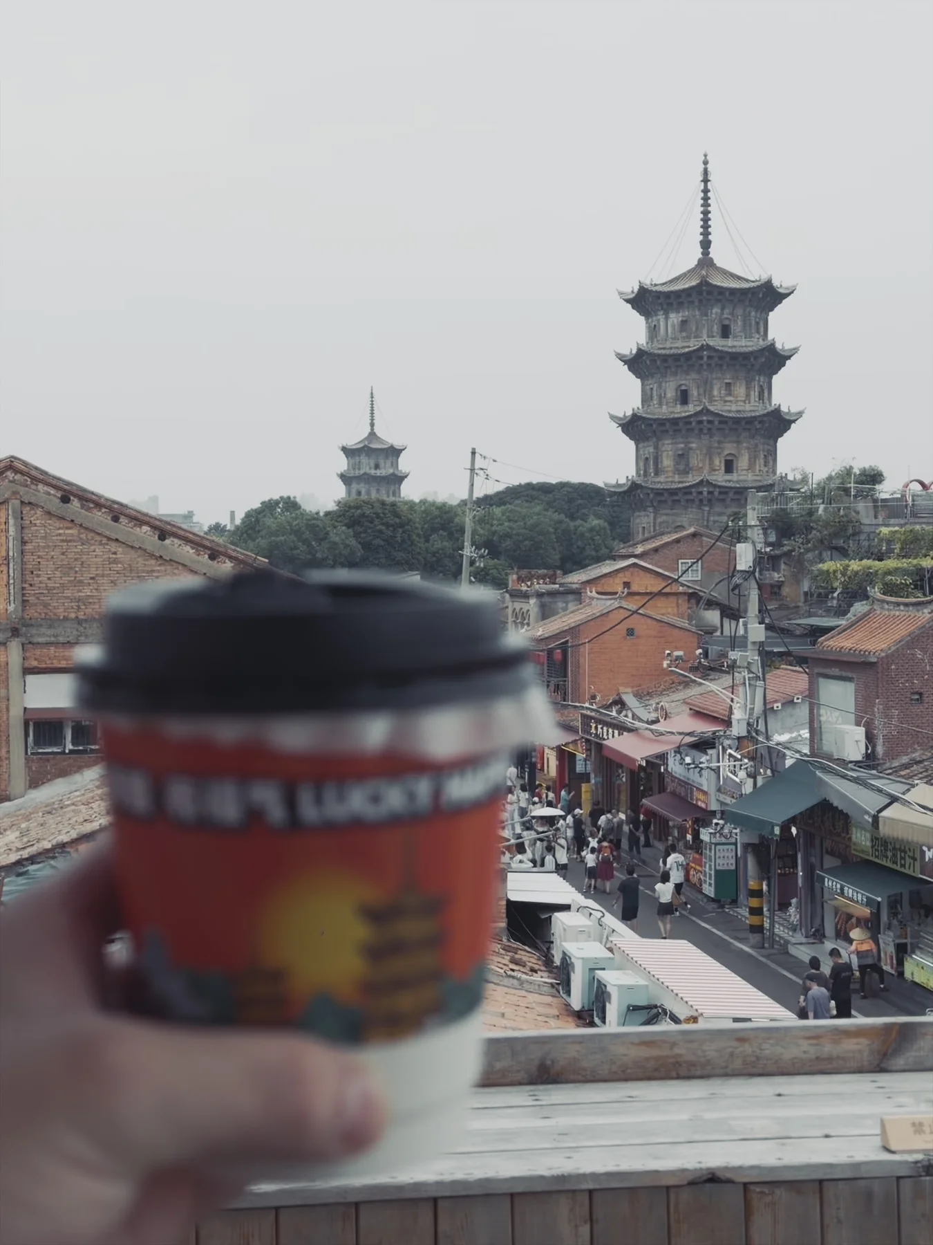 An out-of-focus coffee cup in the foreground, with Kaiyuan Temple’s twin pagodas rising beyond Quanzhou’s red-tiled roofs under a pale evening sky