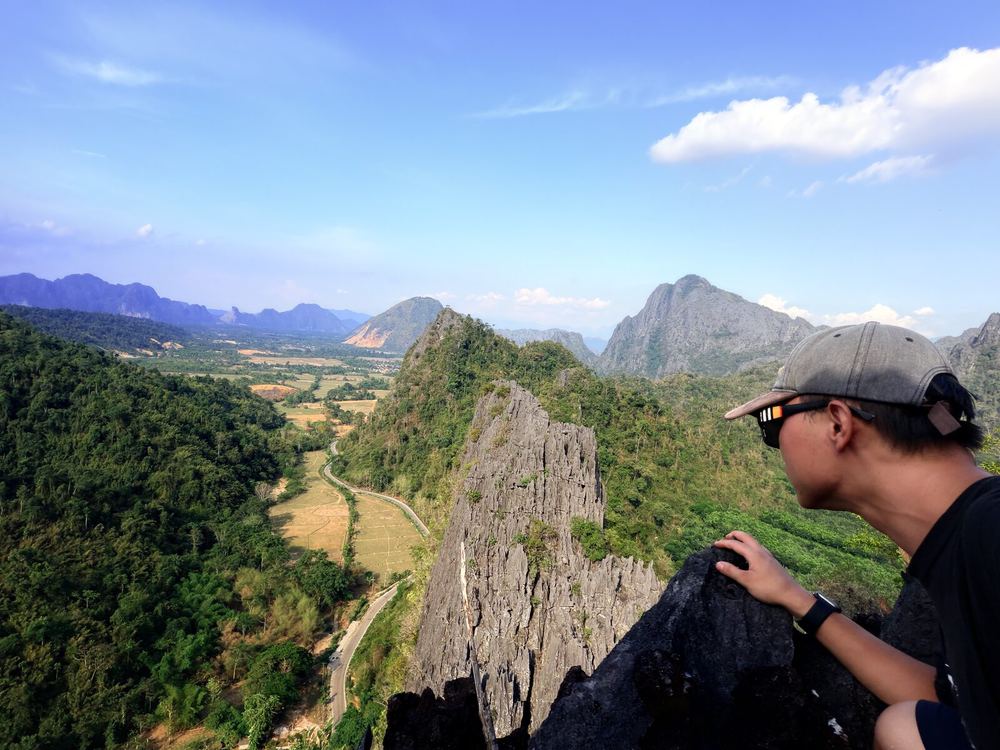 Selfie at a cliff edge on a Vang Vieng summit: the switchback road below, the karst range stretching out in the distance. In that moment you can clearly feel the body&rsquo;s settled certainty on the edge