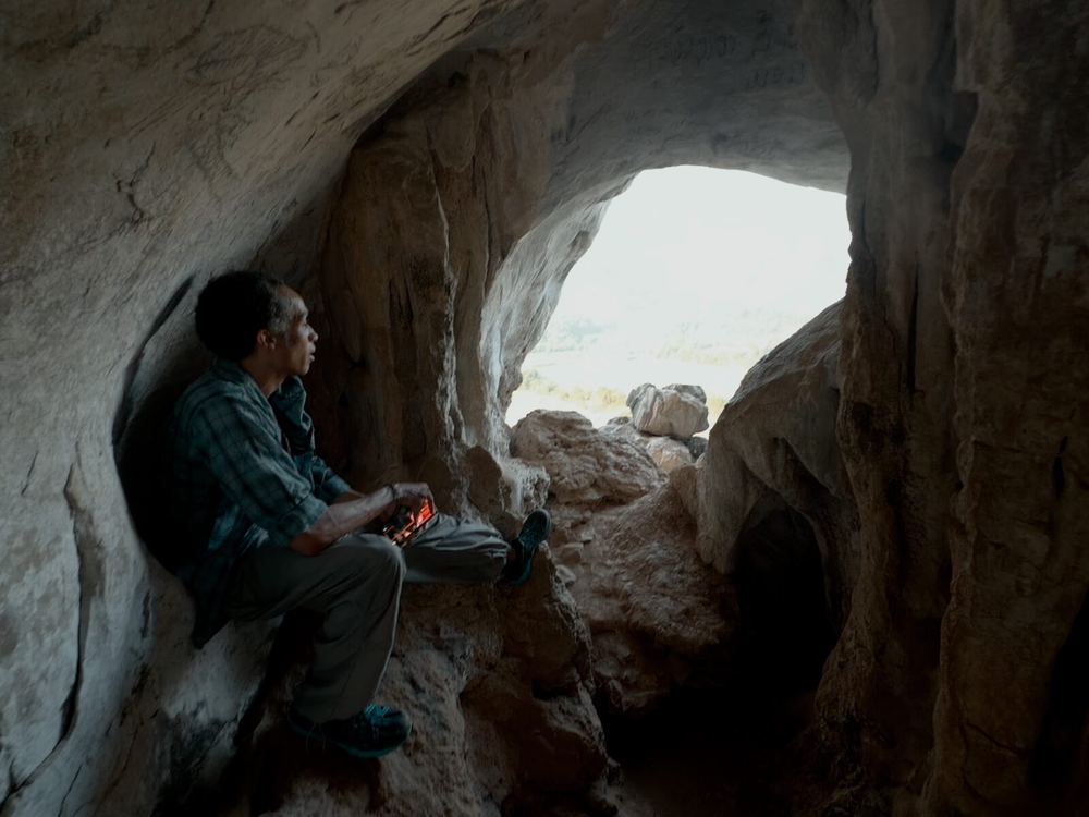 A local Laotian man sitting at the mouth of a cave, looking out toward the light, a small red flame in his hand — as if he were keeping a world that gets to be slow on its own terms
