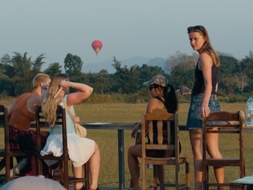 Dusk in Vang Vieng — a hot-air balloon drifts above the rice paddies in the distance, a few travelers sitting at a wooden table. One of the last frames before I left, and the very frame that floats up later, every time I think back on the word &ldquo;reluctance&rdquo;